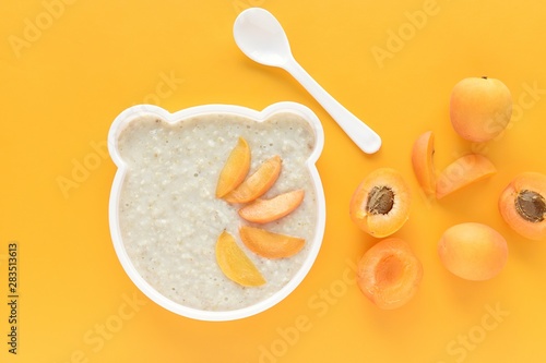 Oatmeal porridge with apricot for kids, funny bear shape plate, baby food, yellow background.