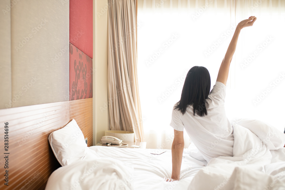 Woman stretching in bed after waking up, back view. Woman sitting near the big white window while stretching on bed after waking up with sunrise at morning, back view. Woman stretching in bed after waking up, back view. Woman sitting near the big white window while stretching on bed after waking up with sunrise at morning, back view.