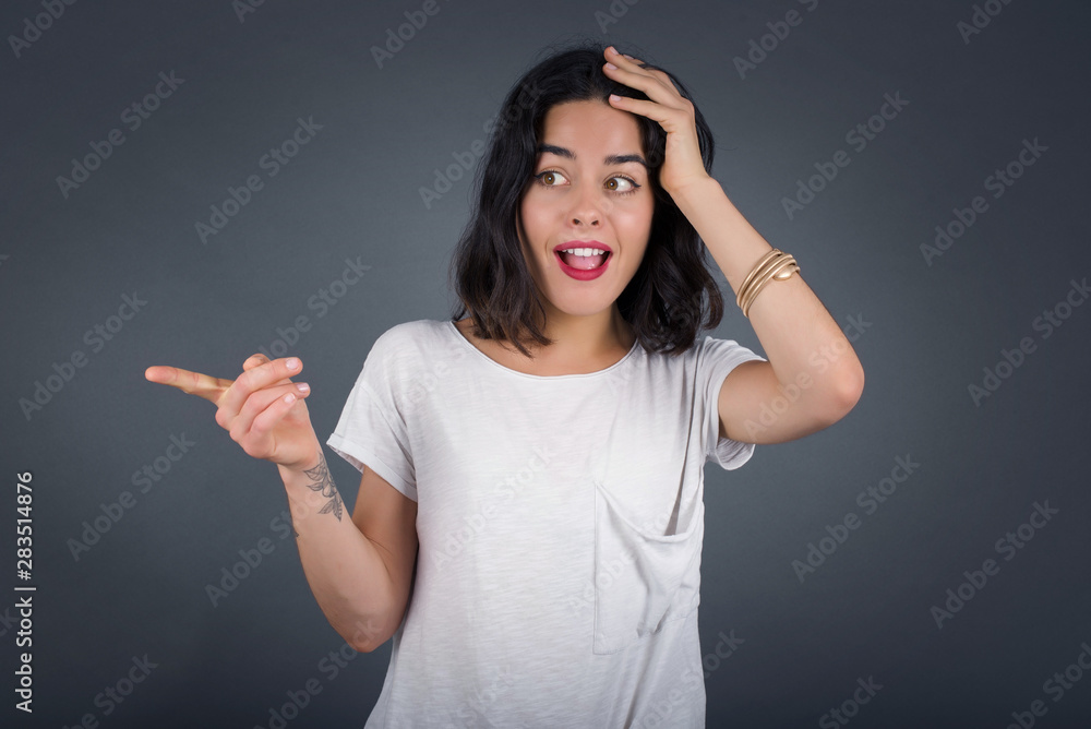 Portrait of young woman with shocked facial expression, showing something amazing on blank space, one hand on her head and pointing with forefinger. OMG concept. Portrait of young woman with shocked facial expression, showing something amazing on blank space, one hand on her head and pointing with forefinger. OMG concept.