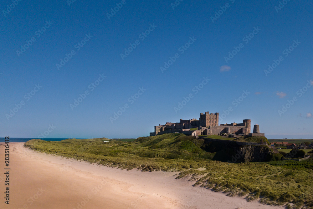 Fototapeta premium Aerial overhead view of Bamburgh Castle in the beautiful quant old Northumbrian village. Drone shot view across the sand dunes and beach set against a brilliant blue sky.