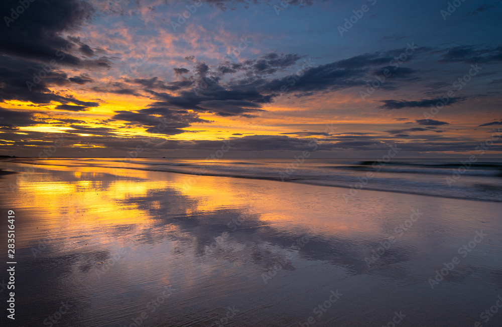 Coastline beach sunset on the shore at Bamburgh, Northumberland in north east England. Incoming tide on the sand providing reflections and symmetry to the colourful and dramatic skies at dusk. Coastline beach sunset on the shore at Bamburgh, Northumberland in north east England. Incoming tide on the sand providing reflections and symmetry to the colourful and dramatic skies at dusk.