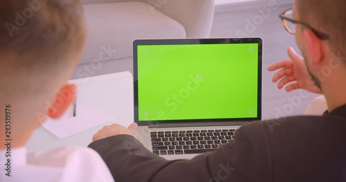 Closeup back view portrait of two successful caucasian businessmen using laptop with green chroma key screen in the office indoors