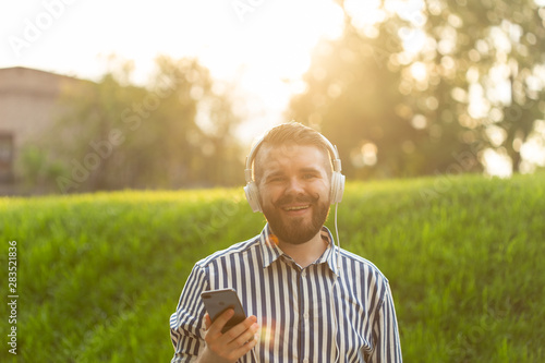 Positive young male hipster listens to your favorite online radio using smartphone headphones and high-speed wireless internet in the park on a sunny warm summer day.