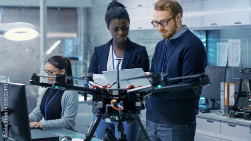 Caucasian Male and Black Female Engineers Working on a Drone Project with Help of Laptop and Taking Notes. He Works in a Bright Modern High-Tech Laboratory.