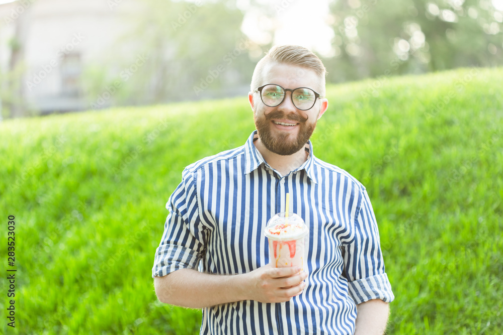 Positive young hipster guy in glasses and stylish beard is drinking a milkshake with a straw while standing outside against a park. The concept of lovers of light sweet drinks. Positive young hipster guy in glasses and stylish beard is drinking a milkshake with a straw while standing outside against a park. The concept of lovers of light sweet drinks.