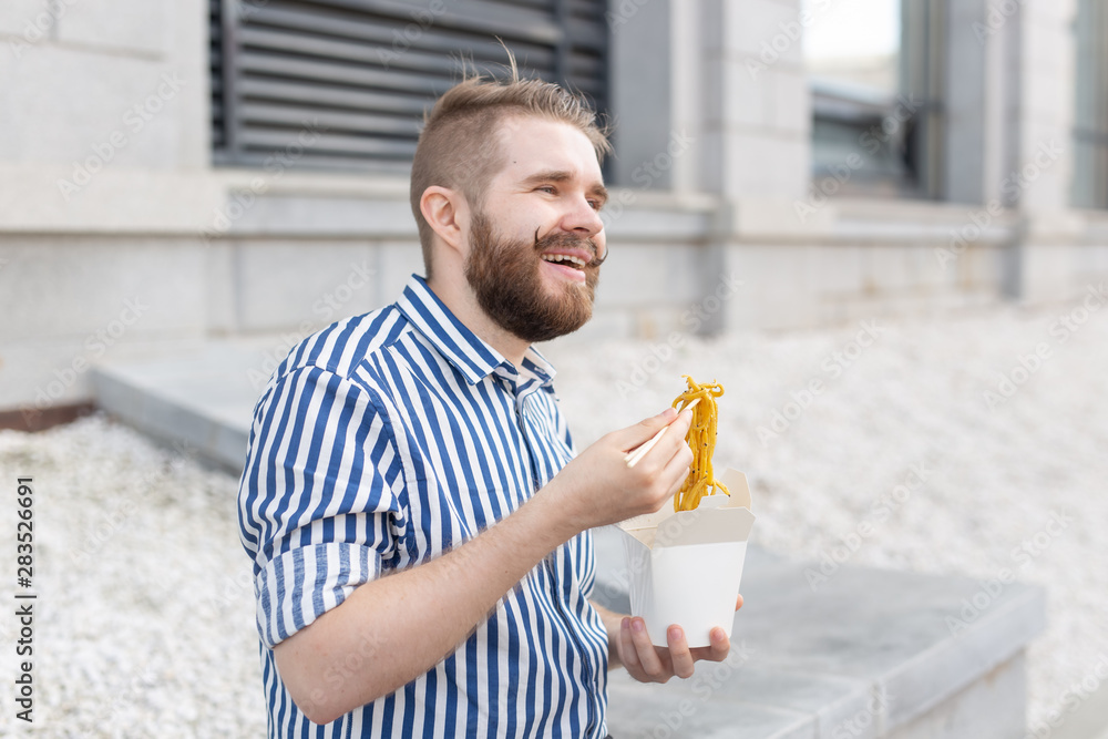 Close-up of an amusing young hipster guy eating chinese noodles with wooden chopsticks sitting in a park outside on a warm summer day. The concept of rest and snacking on the street. Close-up of an amusing young hipster guy eating chinese noodles with wooden chopsticks sitting in a park outside on a warm summer day. The concept of rest and snacking on the street.