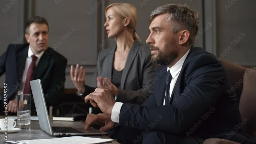 Medium shot of male and female corporate executives in business formal attire sitting together in meeting room and participating in negotiations, man working on laptop and woman talking and gesturing Medium shot of male and female corporate executives in business formal attire sitting together in meeting room and participating in negotiations, man working on laptop and woman talking and gesturing
