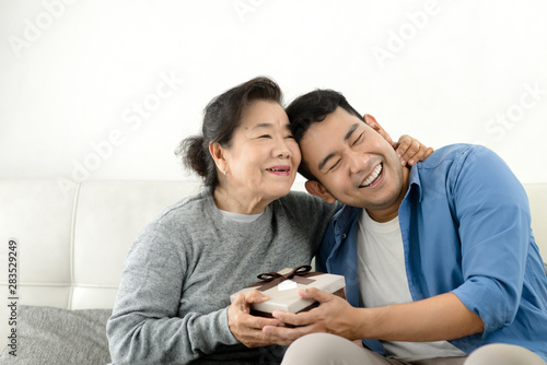 Happy Asian man giving gift box to his mother for Mother's Day celebration.