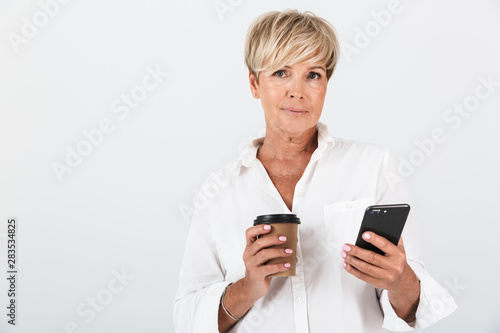 Portrait of attractive adult woman with short blond hair holding cellphone and takeaway coffee cup