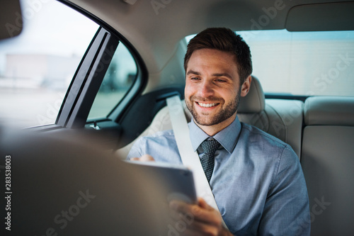 Businessman with tablet sitting on back seats in car, working.