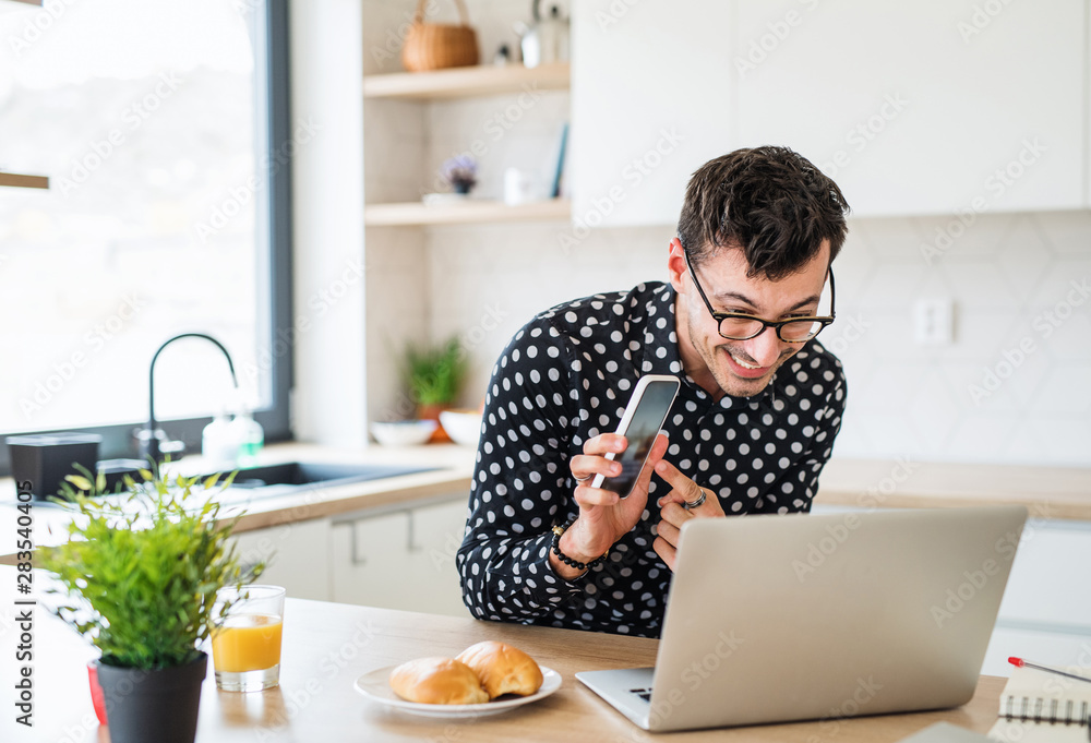 Young man with laptop and smartphone sitting in kitchen, a home office concept. Young man with laptop and smartphone sitting in kitchen, a home office concept.