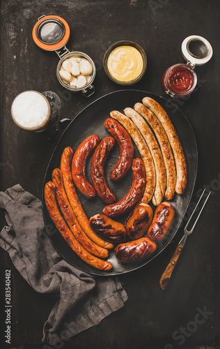 Flat-lay of Octoberfest party dinner table concept with grilled veal and pork sausages, sauce, pickled vaggies and beer in glass over dark background, top view