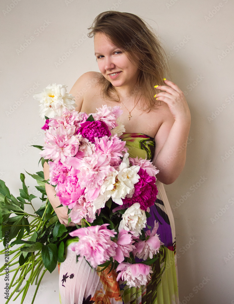 young attractive girl in dress with bouquet of peonies is standing with one hand near hair, looking straight and smiling happy on white wall background, romantic concept, free space young attractive girl in dress with bouquet of peonies is standing with one hand near hair, looking straight and smiling happy on white wall background, romantic concept, free space