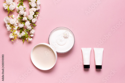 Beauty Spa concept. Opened plastic container with cream, cosmetic bottle containers, spring White flowers on pink background Flat lay top view. Herbal dermatology cosmetic hygienic cream organic
