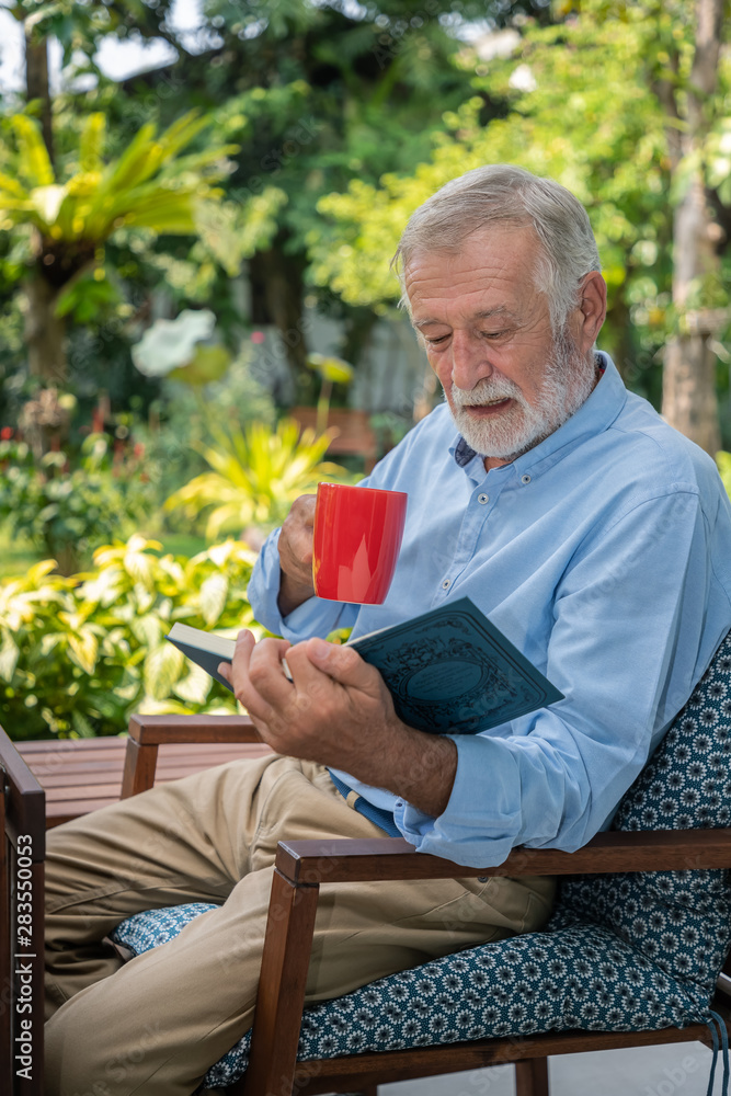 Senior elderly man reading book holding mug of coffee in garden