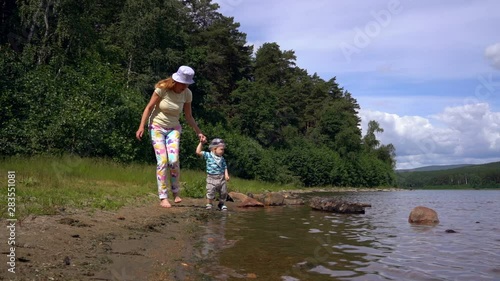 Mom and her little son walk along the shore of a forest lake on a summer sunny day against the background of the forest and blue sky. Mom holds the baby's hand, sometimes they stop and touch the water