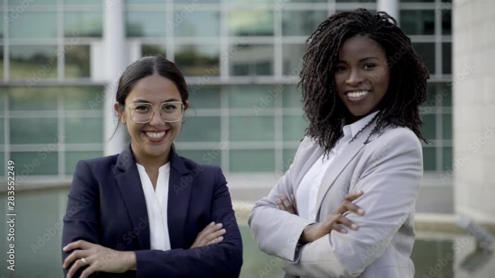 Two smiling colleagues standing with crossed arms on street. Cheerful young confident employees wearing formal suits looking at camera. Business confidence concept Two smiling colleagues standing with crossed arms on street. Cheerful young confident employees wearing formal suits looking at camera. Business confidence concept