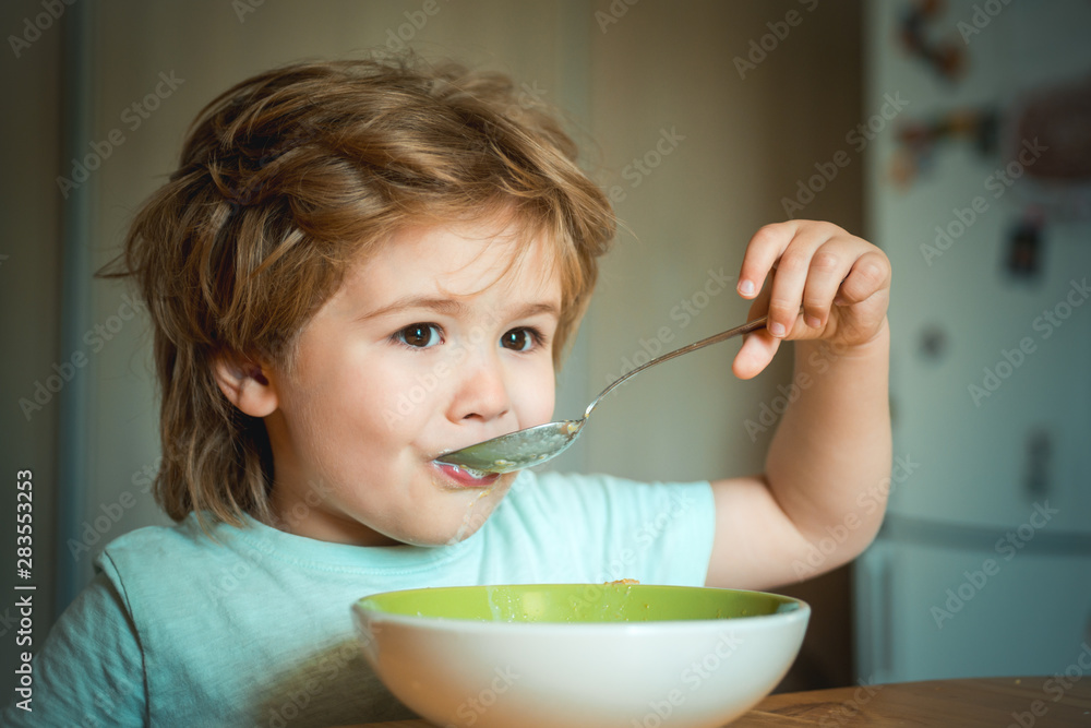 Laughing cute child baby boy sitting in highchair and eating on blurred background. Cheerful baby child eats food itself with spoon. Parenthood. Little boy sitting at the table and eating milk snack. Laughing cute child baby boy sitting in highchair and eating on blurred background. Cheerful baby child eats food itself with spoon. Parenthood. Little boy sitting at the table and eating milk snack.
