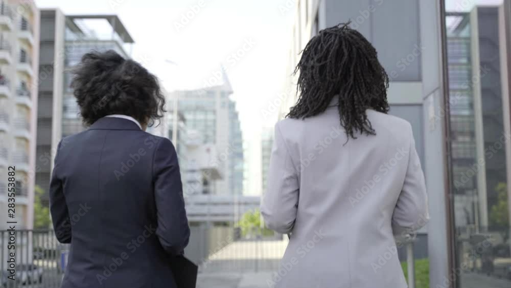Back view of businesswomen walking on street. Rear view of professional multiethnic young female colleagues holding folders and talking while walking outside modern office building. Business concept Back view of businesswomen walking on street. Rear view of professional multiethnic young female colleagues holding folders and talking while walking outside modern office building. Business concept