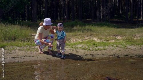 Mom and her little son play with pebbles near the water on the shore of a forest lake. Mother and her baby together this summer weekend