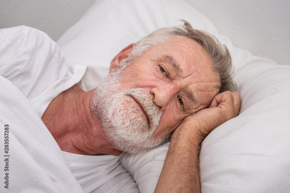 Senior elderly man on bed eye open sleeplessness and worry, with white blanket in bedroom Senior elderly man on bed eye open sleeplessness and worry, with white blanket in bedroom