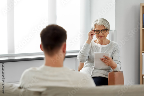 psychology, mental therapy and people concept - senior woman psychologist with clipboard and young man patient at psychotherapy session