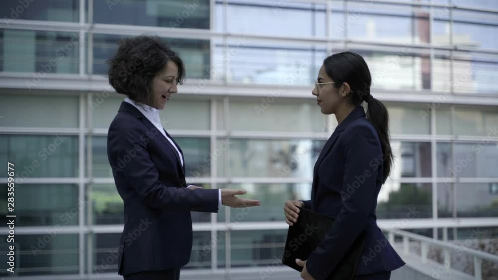 Cheerful businesswomen talking outdoor. Side view of professional young businesswomen shaking hands and talking outside modern office building. Business concept Cheerful businesswomen talking outdoor. Side view of professional young businesswomen shaking hands and talking outside modern office building. Business concept