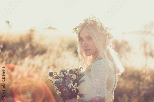 Beautiful young woman portrait in a white dress in boho style with a floral wreath in the summer in the field. Selective soft focus.