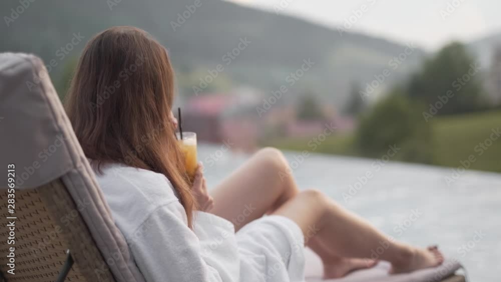 Static shot of a beautiful sexy lady sitting on a sunbed wearing a beach robe and sipping on a drink with a pool and greenery blurred in the background Static shot of a beautiful sexy lady sitting on a sunbed wearing a beach robe and sipping on a drink with a pool and greenery blurred in the background
