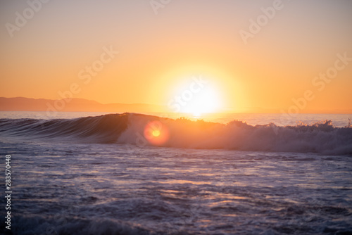 Landscape shot of waves crashing with bright sun rising over the horizon in background