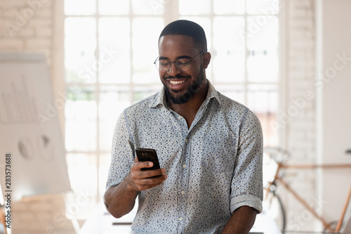 Happy black businessman holding smartphone using apps standing in office