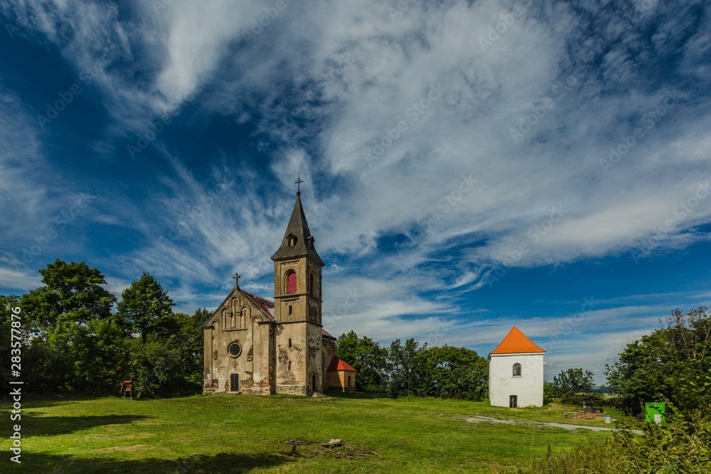 Krasikov, Kokasice / Czech Republic - August 9 2019: View of the church of Mary Magdalene and a bell tower. Sunny summer day. Green grass, blue sky with white clouds.