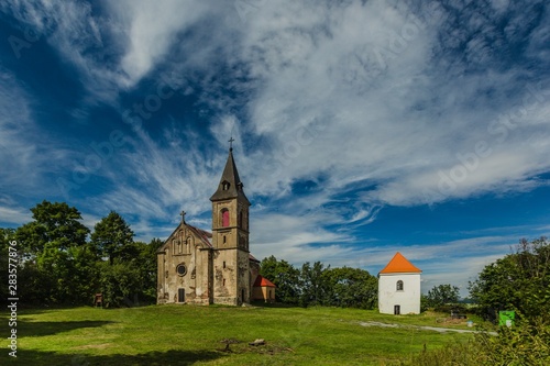 Krasikov, Kokasice / Czech Republic - August 9 2019: View of the church of Mary Magdalene and a bell tower. Sunny summer day. Green grass, blue sky with white clouds.