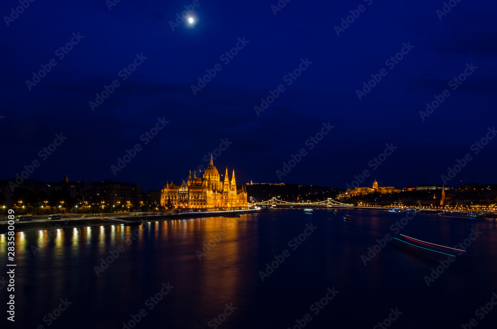 Nightscape city view of Budapest parliament under the cloud covered moon in the sky and water reflection