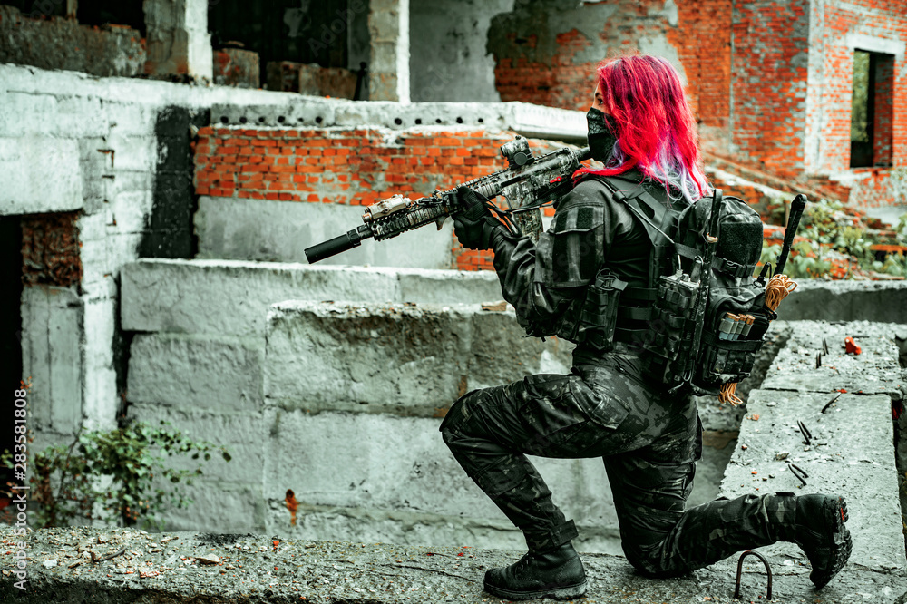 Fototapeta premium Airsoft red-hair woman in uniform with machine gun standing on knee. Soldier on ruine. side view