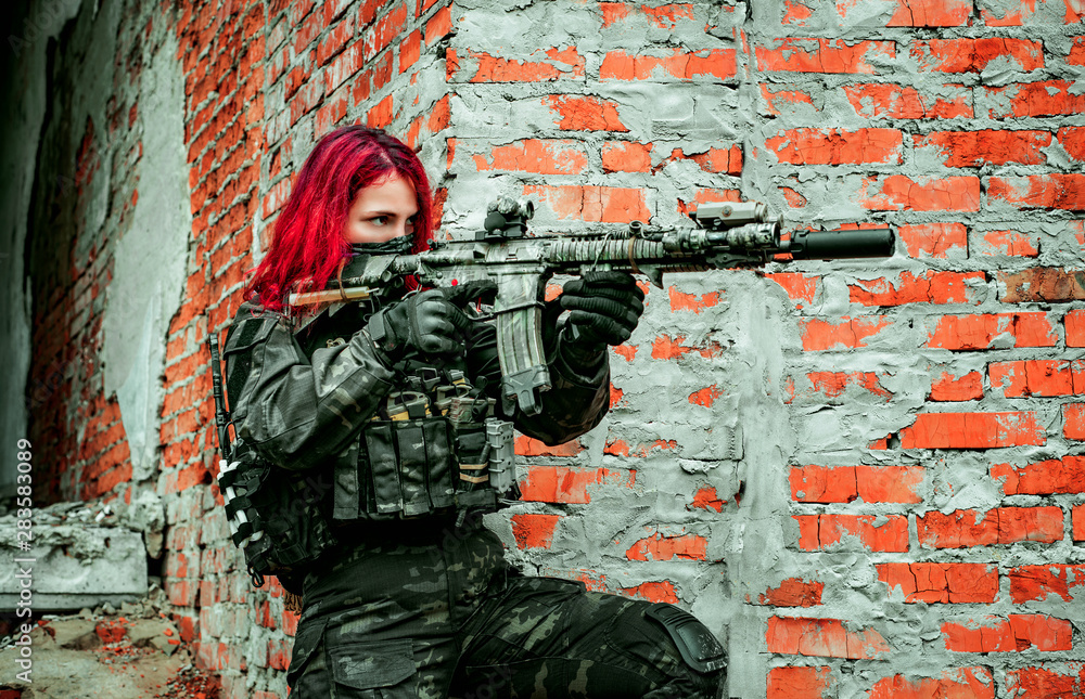 Fototapeta premium Airsoft red-hair woman in uniform with machine gun beside brick wall. Soldier aims at the sight on the ruins. Side view