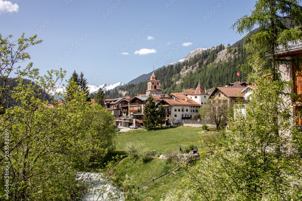 View of a village in the swiss alps with meadows, mountains, cows, church and beautiful houses in spring at sunshine