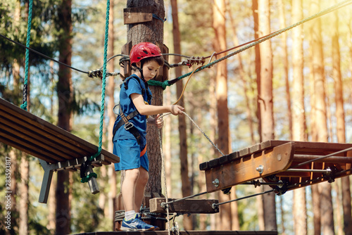 Boy enjoying activity in climbing adventure park at sunny summer day. Kid climbing in rope playground structure. Safe climbing with helmet insurance. Child in forest adventure park, extreme sport.