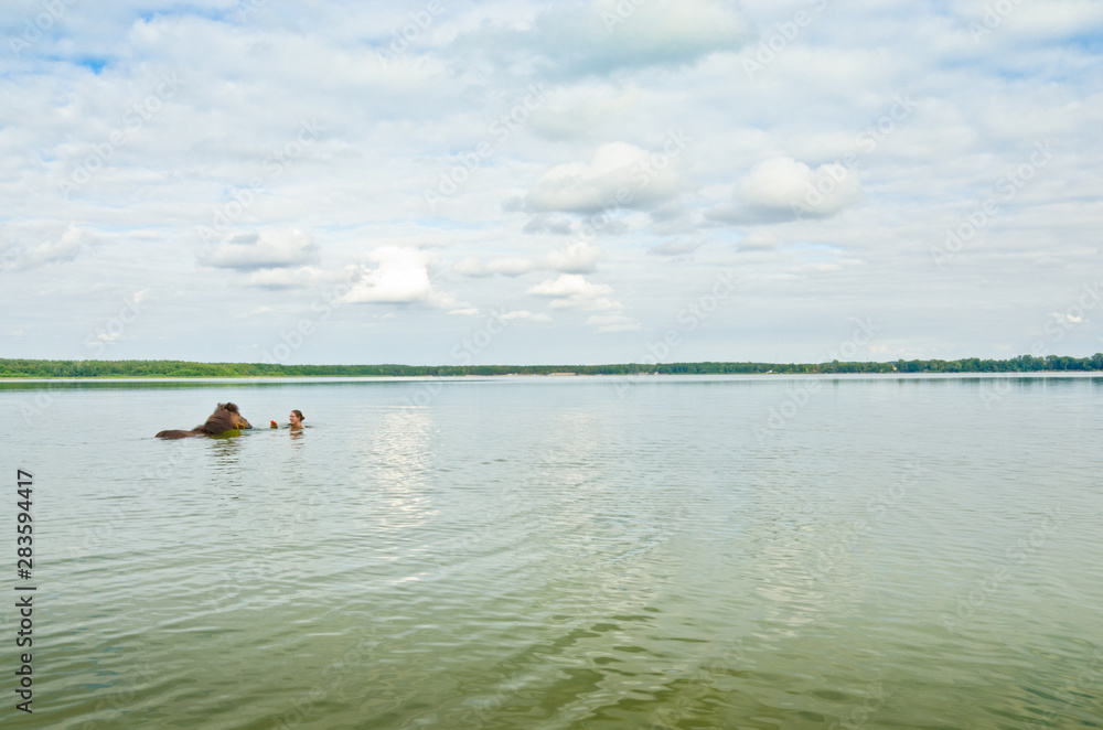 Eine Frau badet mit einem braunen Pferd weit draußen in einem See und lockt es mit einem ins tiefe Wasser 