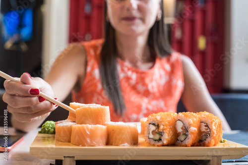 Hand of a girl in a red dress with red nails, takes fish sushi roll to chopsticks. Roll s stand on a wooden stand. Close up photo