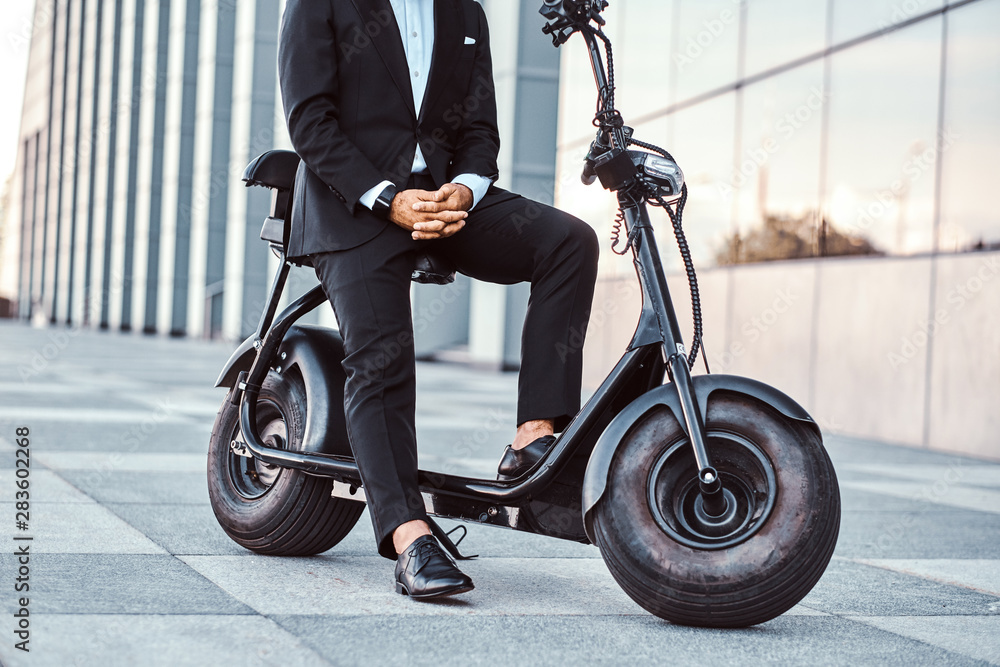 Elegant man in black suit is sitting on his electro scooter while posing for photographer.