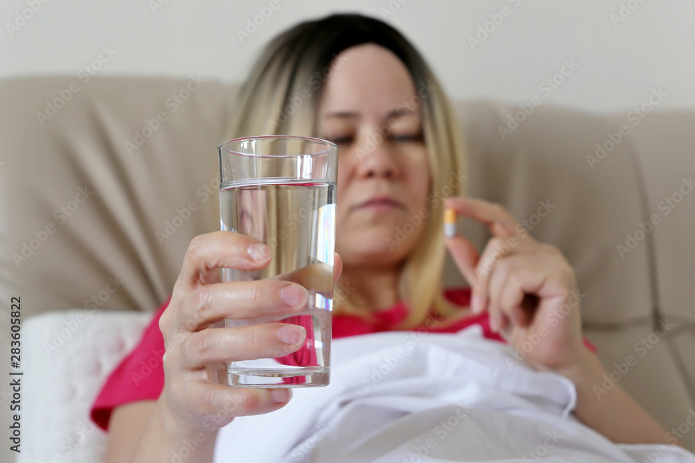 Woman taking a pill in a bed holding a water glass. Medication in capsule, concept of antibiotic, vitamin, cold and flu Woman taking a pill in a bed holding a water glass. Medication in capsule, concept of antibiotic, vitamin, cold and flu