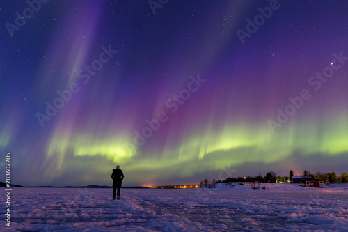 Person watching a breath-taking display of colorful northern lights on frozen lake Inari, Finland.