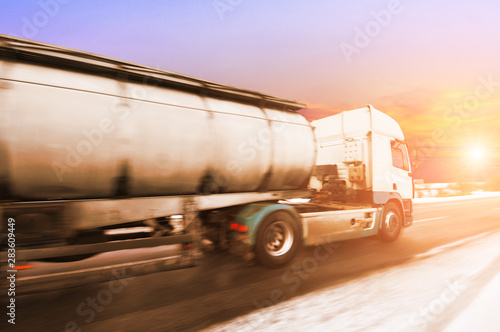 Big metal fuel tanker truck in motion shipping fuel on the winter countryside road with snow against sky with sunset