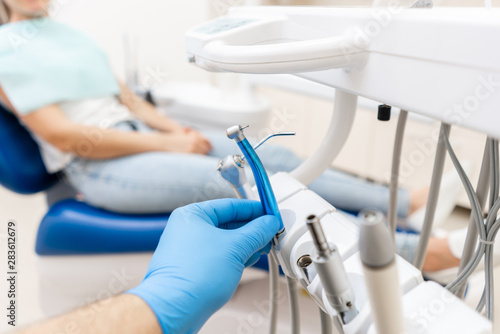 Close-up hand of dentist in the glove holds dental high speed turbine. The patient in blue chair at the background. Office where dentist conducts inspection and concludes.