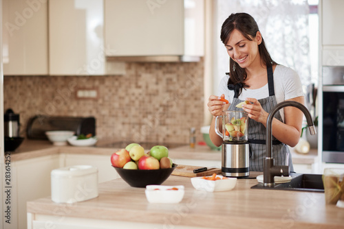 Beautiful Caucasian smiling woman in apron putting vegetables and fruits in blender while standing in kitchen.