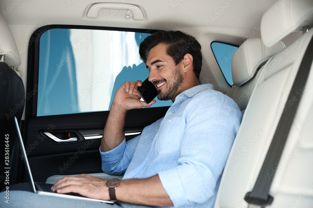 Attractive young man working with laptop and talking on phone in luxury car