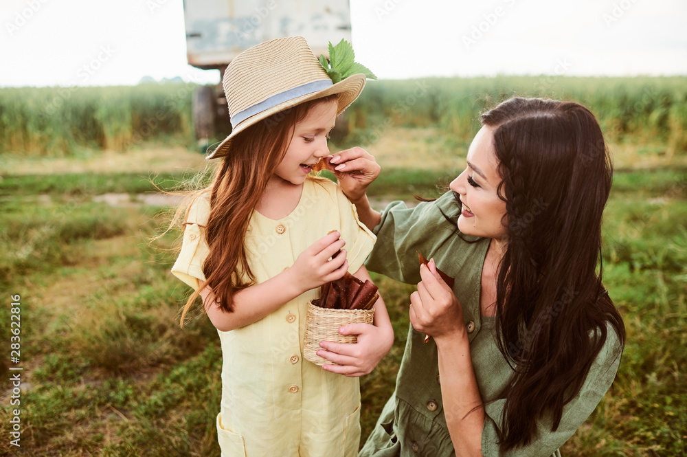 Fototapeta premium Beautiful young caucasian mother with her daughter in a linen dress with a basket of strawberries snacks gathers a new crop and has fun in the green field