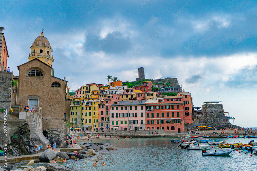 The marina of Vernazza in Liguria, Cinque Terre, Italy. Boats and colorful historical houses and people swimming on the Mediterranean sea. Blue sky on the European summer.