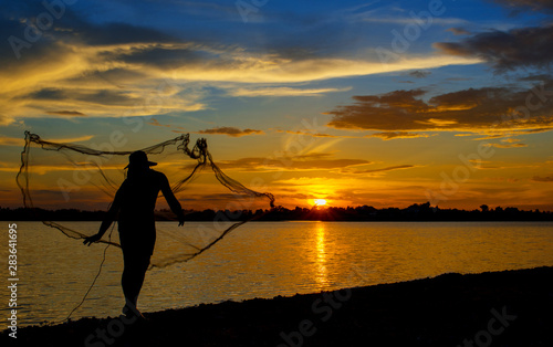 Silhouette of Thai Fisherman catching the fish in twilight, Beautiful Sunset in the sky with sky blue and orange light of the sun through the clouds in the sky, Orange and red dramatic colors.